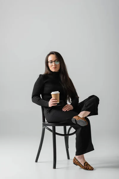 Full length of asian woman in black outfit and glasses holding paper cup while sitting on chair on grey — Fotografia de Stock