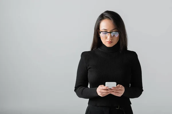 Young asian woman in glasses and black turtleneck texting on smartphone isolated on grey - foto de stock