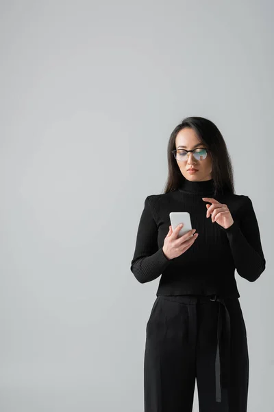 Brunette asian woman in glasses and black turtleneck using smartphone isolated on grey — Stock Photo