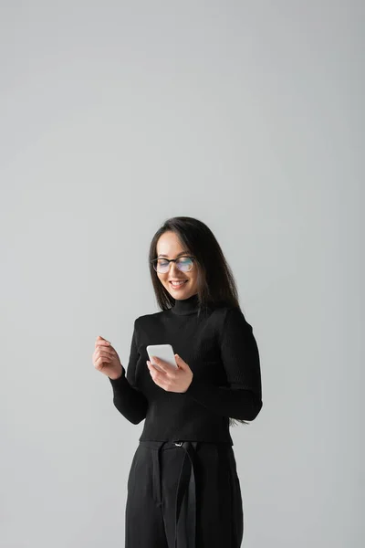 Cheerful asian woman in glasses and black turtleneck using smartphone isolated on grey — Stock Photo