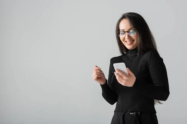 Happy asian woman in glasses and black turtleneck using smartphone isolated on grey — Stock Photo