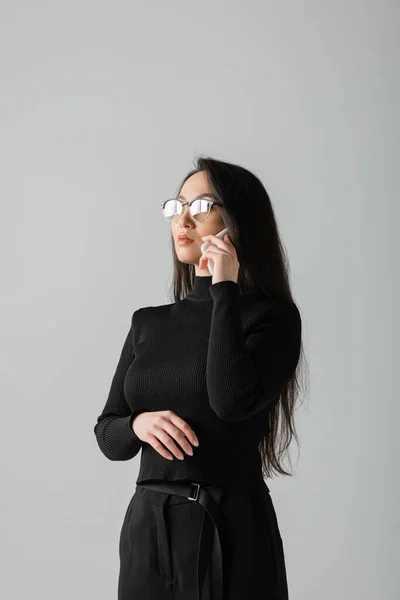 Young asian woman in glasses and black turtleneck talking on smartphone isolated on grey — стоковое фото