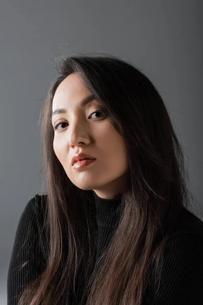 Portrait of young and asian woman in black turtleneck looking at camera isolated on dark grey — стоковое фото
