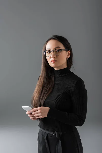 Brunette asian woman in black turtleneck and glasses holding smartphone on dark grey — Fotografia de Stock