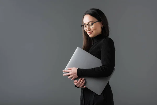 Cheerful asian woman in black turtleneck and glasses holding laptop isolated on dark grey — стоковое фото