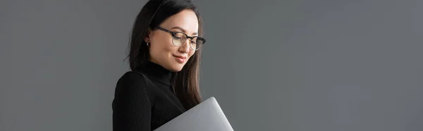 Cheerful asian woman in black turtleneck and glasses holding laptop isolated on dark grey, banner — Fotografia de Stock
