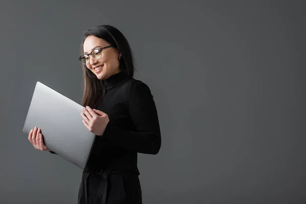 Positive asian woman in black turtleneck and glasses holding laptop isolated on dark grey — стоковое фото