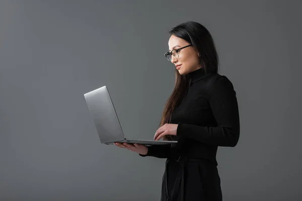 Pretty asian woman in black turtleneck and glasses using laptop isolated on dark grey — Fotografia de Stock