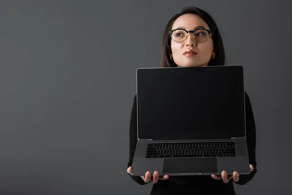 Pretty asian woman in black turtleneck and glasses holding laptop with blank screen isolated on dark grey - foto de stock