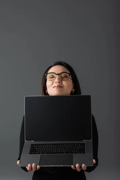 Pleased asian woman in black turtleneck and glasses holding laptop with blank screen isolated on dark grey - foto de stock