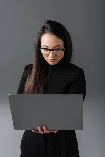 Brunette asian woman in black turtleneck and glasses holding laptop on dark grey - foto de stock