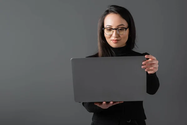 Smiling asian woman in black turtleneck and glasses holding laptop isolated on dark grey — стоковое фото