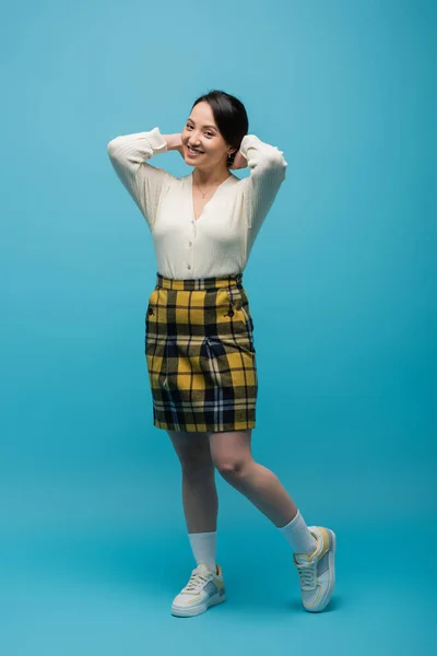 Full length of happy asian woman in checkered skirt and sneakers standing on blue — Stock Photo