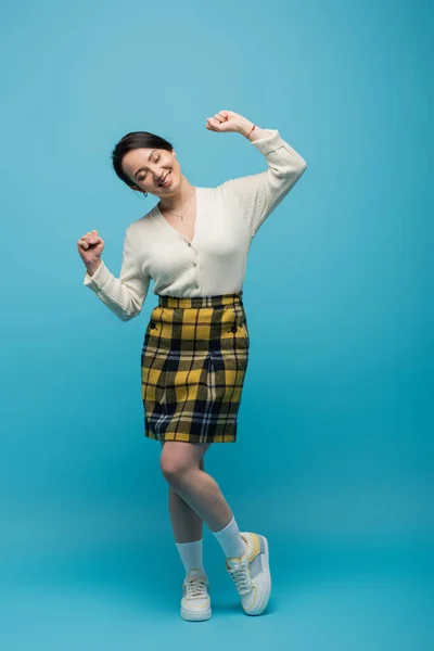 Full length of cheerful asian woman in checkered skirt and sneakers gesturing on blue — Stock Photo