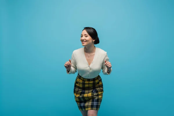 Cheerful asian woman in plaid skirt smiling while posing isolated on blue — Photo de stock