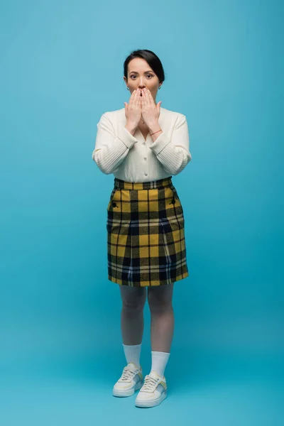 Full length of shocked asian woman in checkered skirt and sneakers covering mouth on blue — Stock Photo