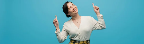 Cheerful asian woman in plaid skirt smiling while pointing up with fingers isolated on blue, banner — стоковое фото