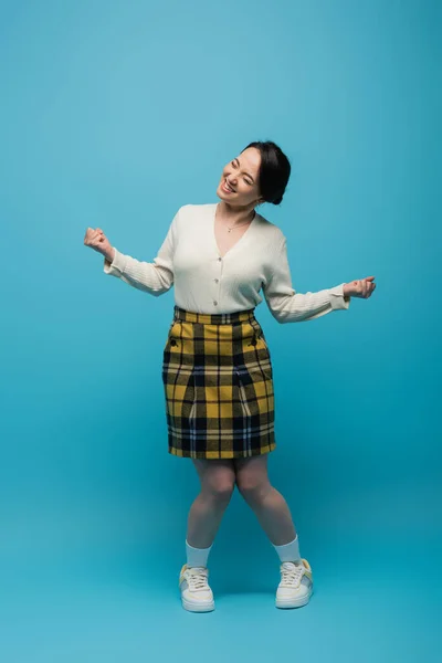 Full length of joyful asian woman in checkered skirt and white sneakers standing on blue - foto de stock