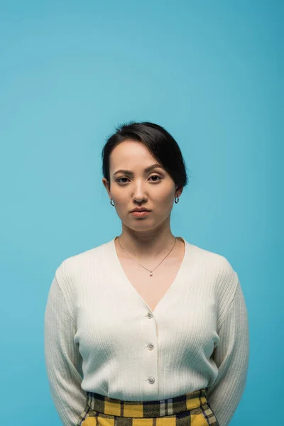 Serious asian woman in cardigan looking at camera  isolated on blue - foto de stock