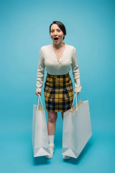 Full length of excited asian woman holding shopping bags on blue background - foto de stock
