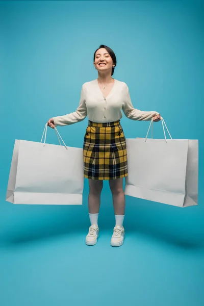Positive asian woman holding shopping bags while standing on blue background - foto de stock