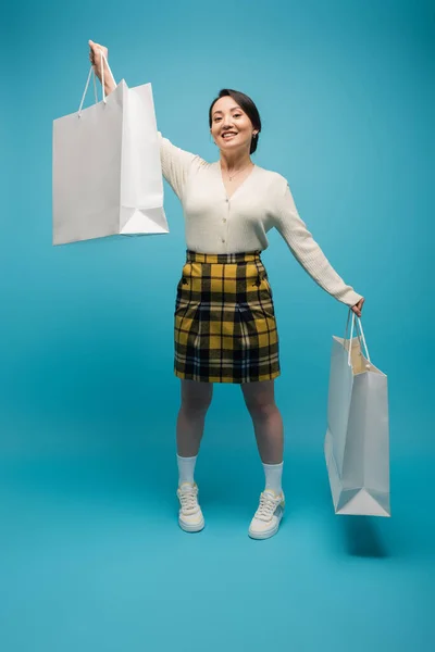 Full length smiling asian woman in plaid skirt and cardigan holding shopping bags on blue background - foto de stock