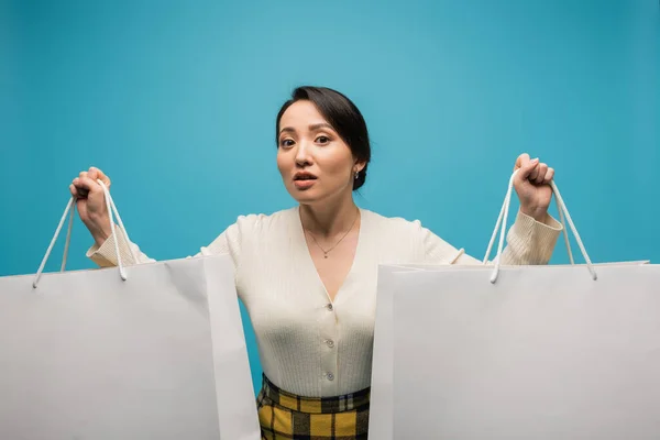 Asian woman holding shopping bags and looking at camera isolated on blue — Stock Photo