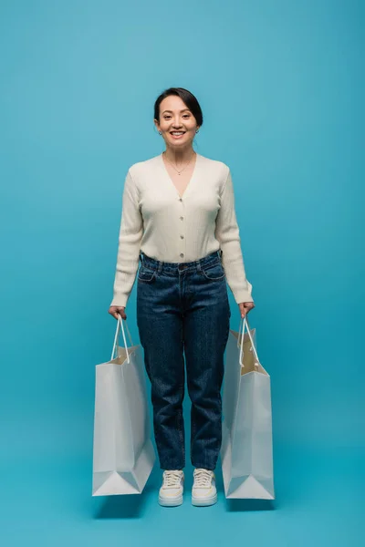 Full length of cheerful asian woman in jeans and cardigan holding shopping bags on blue background - foto de stock