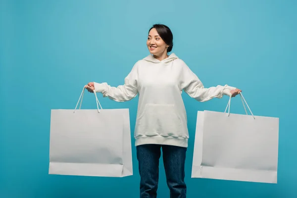 Positive asian woman in hoodie and jeans holding shopping bags isolated on blue - foto de stock