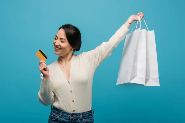 Cheerful asian woman holding credit card and shopping bags isolated on blue — стоковое фото