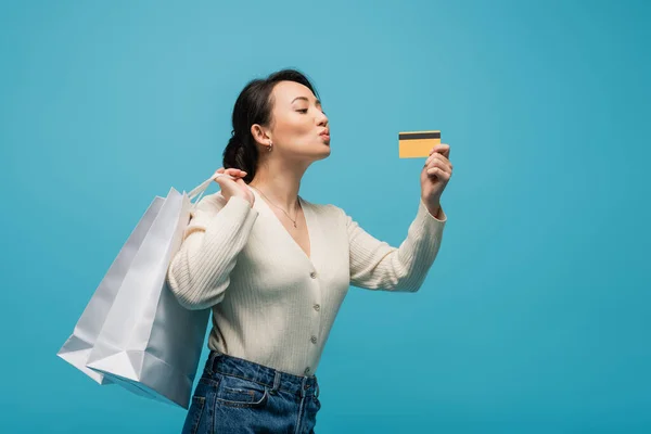 Young asian model pouting lips while holding credit card and shopping bags isolated on blue — Photo de stock