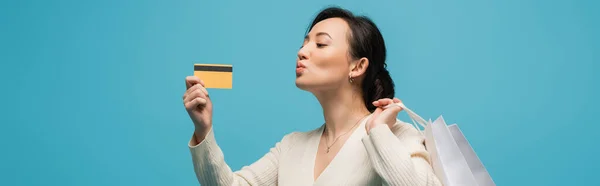 Asian woman holding shopping bags and pouting lips while looking at credit card isolated on blue, banner — стоковое фото