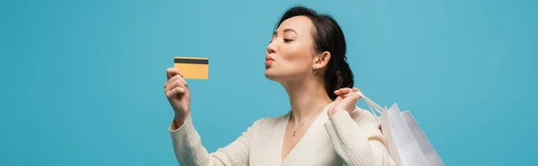 Asian woman holding shopping bags and pouting lips while looking at credit card isolated on blue, banner — Stock Photo