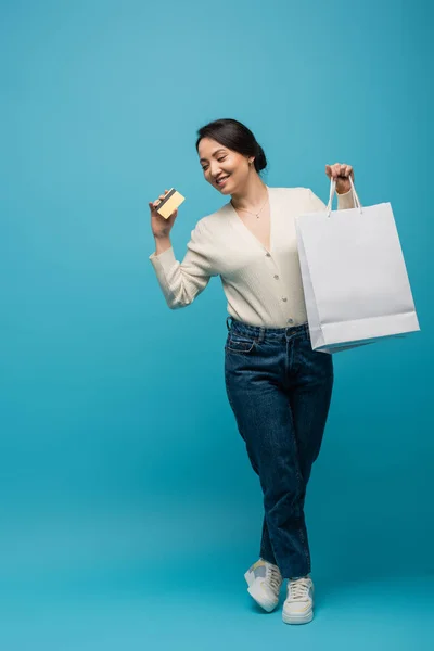 Cheerful asian woman in cardigan and jeans holding shopping bags and credit card on blue background — Fotografia de Stock