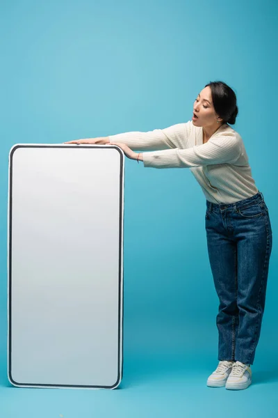 Shocked asian woman in jeans and cardigan standing near big smartphone model on blue background — Stock Photo