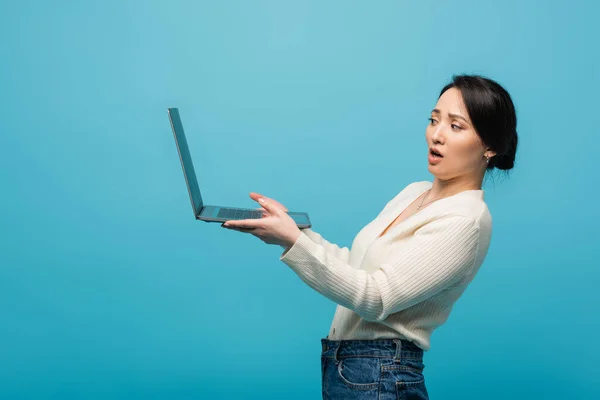 Embarrassed asian woman holding laptop isolated on blue — Stockfoto