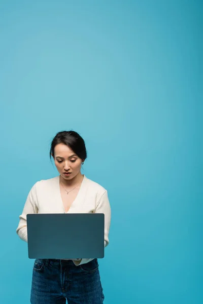 Focused asian freelancer using laptop isolated on blue — стоковое фото