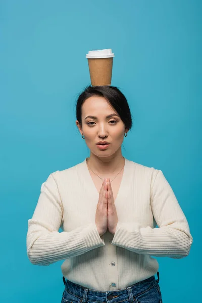 Asian woman with coffee to go on head doing praying hands gesture isolated on blue — Stock Photo