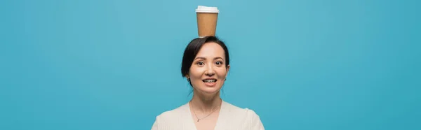 Smiling asian woman holding coffee to go on head isolated on blue, banner — Stock Photo
