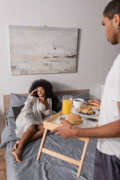 Verschwommene afrikanisch-amerikanische Mann hält Tablett mit Frühstück in der Nähe sexy Freundin auf dem Bett liegend — Stockfoto
