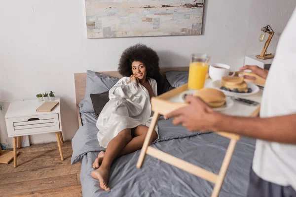Seductive african american woman in silk robe lying on bed near blurred man holding tray with breakfast — Stock Photo