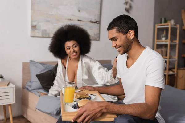 Happy african american man holding tray with pancakes and drinks near smiling brunette woman in silk robe — Stock Photo