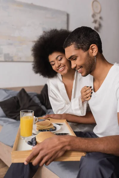 Feliz homem americano africano segurando bandeja com panquecas e bebidas perto de namorada sexy no quarto — Fotografia de Stock