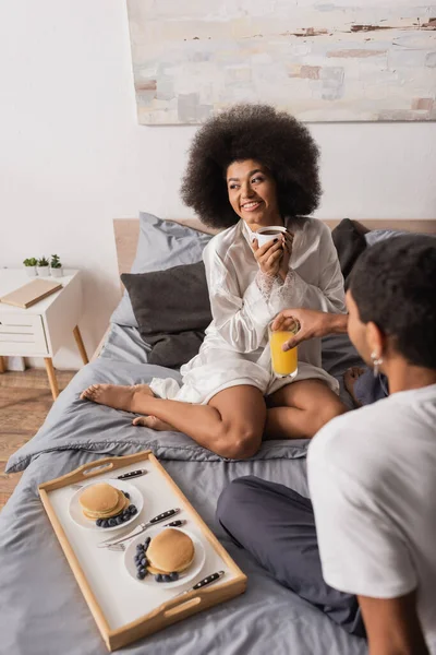 Sexy african american woman in silk robe sitting on bed with orange juice near pancakes with berries and boyfriend with coffee cup — Stock Photo