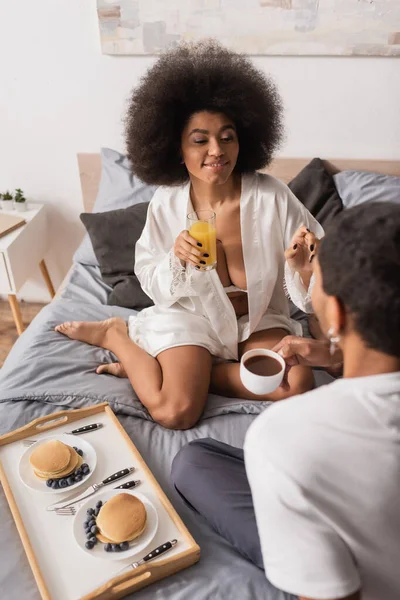 Seductive african american woman in white robe holding orange juice near boyfriend with coffee cup and pancakes on bed — Fotografia de Stock