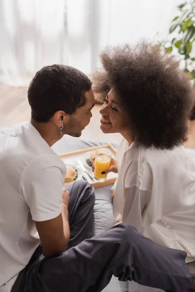 Young and curly african american woman smiling near boyfriend while holding fresh orange juice near breakfast on bed — Stockfoto