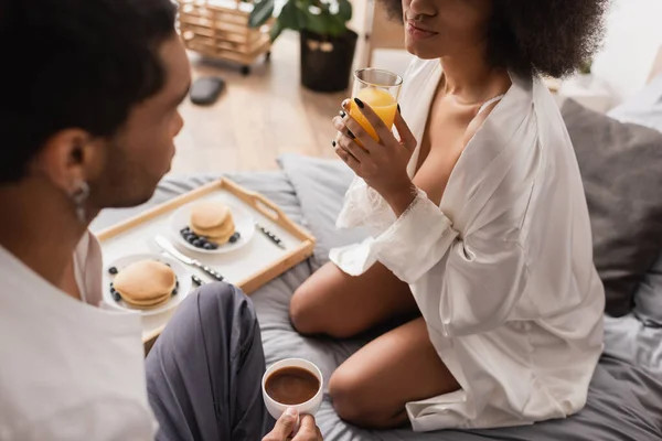 Seductive african american woman sitting with orange juice near delicious breakfast and young boyfriend with coffee cup in bedroom — Stock Photo