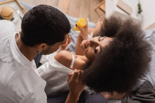 Top view of young african american man undressing sexy girlfriend holding blurred orange juice in bedroom - foto de stock