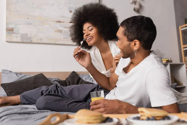 Афроамериканець тримає виделку з чорницею біля веселої дівчини під час сніданку в спальні. — стокове фото