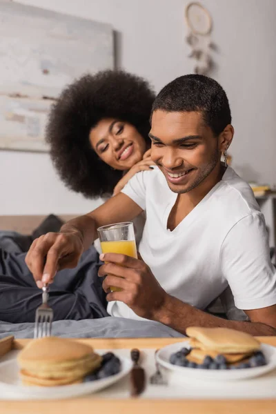 Curly african american woman smiling near boyfriend holding orange juice and fork near blurred pancakes with blueberries — Fotografia de Stock